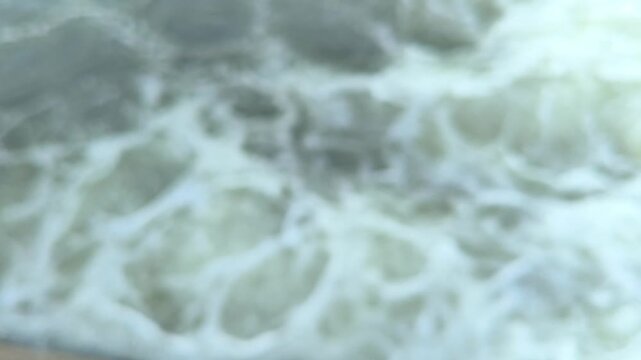 A blurred and defocused view of turbulent water swirling, churning, and producing soft foamy patterns, creating an abstract fluid background with dynamic motion and atmospheric texture.