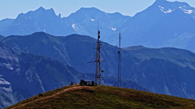 Aerial close-up parallax shot reveals hikers resting by telecom antennas atop grassy mountain crest with jagged Pyrenees peaks at Col d'Aubisque