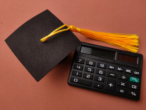 A black graduation cap with a yellow tassel sits beside a black electronic calculator on a brown surface