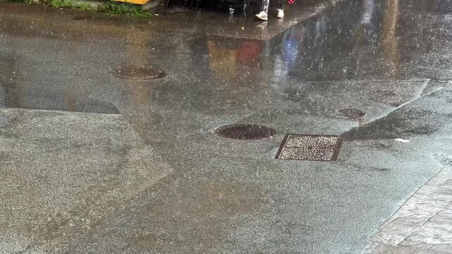 Steady rain pours onto wet asphalt street covered with manhole covers and drains reflecting colorful storefronts nearby