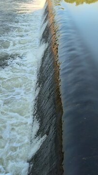 Fast-flowing water rushing over a small dam structure, creating white foam, turbulent currents, and dynamic cascades that highlight the power and movement of flowing water