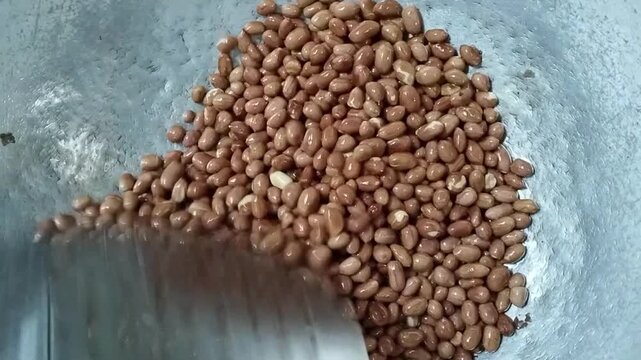 The process of frying peanuts in a concave aluminum wok using a stainless steel slotted spoon or skimmer featuring small perforations.