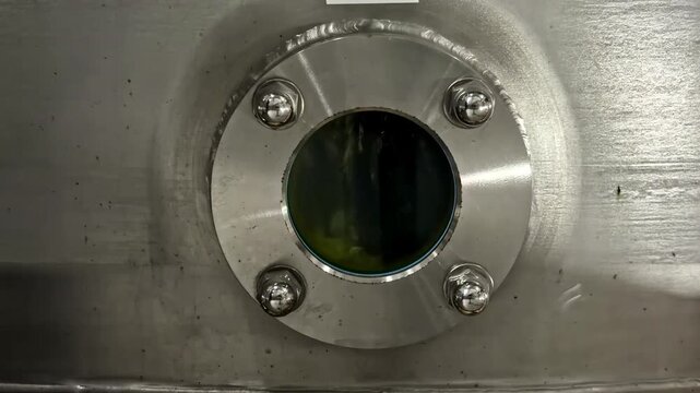 Close-up of stainless steel inspection window on industrial processing tank as fluid sloshes down into vat