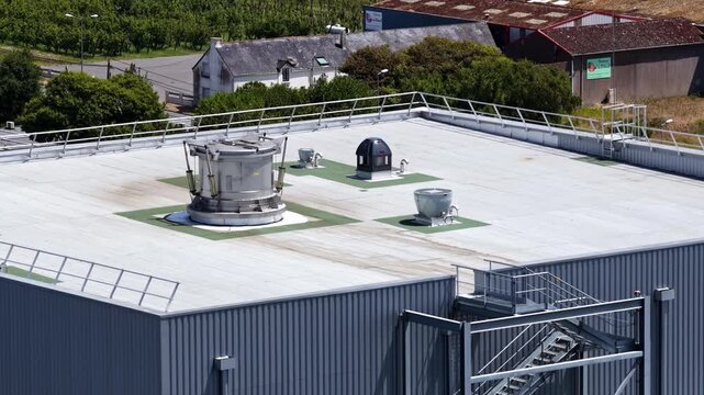 Metal warehouse roof with ventilation equipment and access stairs is filmed by a drone against surrounding green fields.