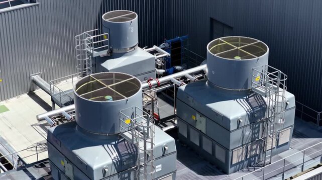 Three large rooftop cooling tower units with ladders and pipework are filmed from above as the drone glides past in bright daylight.