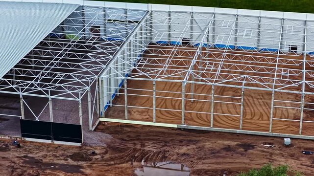 Steel warehouse framework rises over a muddy construction site as the camera moves across the open structure from above.