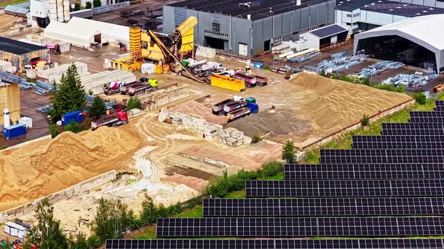 Long rows of solar panels border an active sand quarry and factory yard with heavy equipment on a clear day.