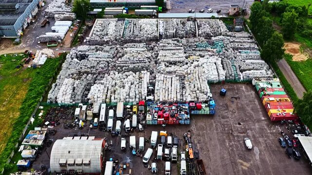Huge plastic recycling yard packed with baled waste, parked trucks, and containers seen from a high drone flyover.