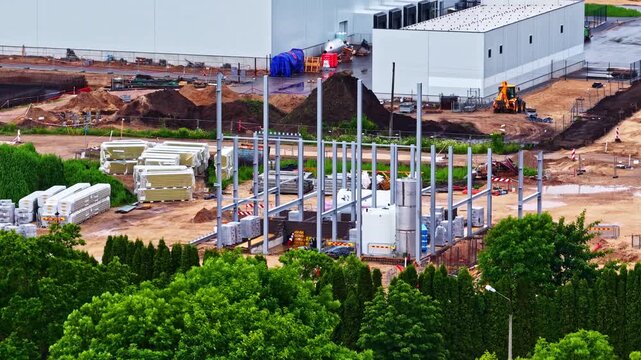 Steel columns rise on a large construction site beside existing factory buildings viewed from an elevated drone perspective.