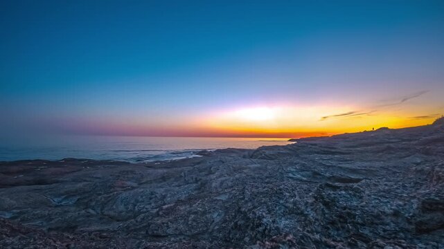 Vivid orange sunset fades into deep blue sky over rugged rocky coastline as gentle waves wash textured rocks along horizon