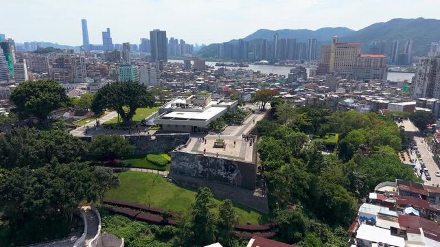 Forward aerial view approaching Mount Fortress and Macau Museum with historic walls, lush gardens and panoramic Macau skyline beside Pearl River