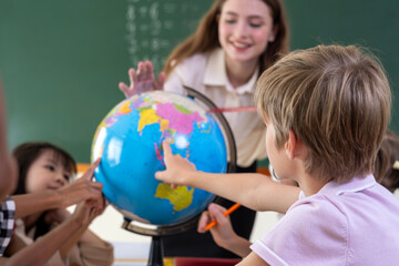 Smiling female teacher guiding elementary students with a world globe. Curious schoolboy pointing at continents on globe during geography class. Young diverse students learning world geography. © unikyluckk