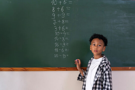 Young schoolboy standing by chalkboard with math equations. African American student holding chalk at blackboard in classroom. Focused elementary student learning mathematics during school lesson.