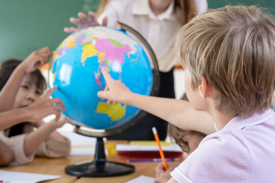 Smiling female teacher guiding elementary students with a world globe. Curious schoolboy pointing at continents on globe during geography class. Young diverse students learning world geography.