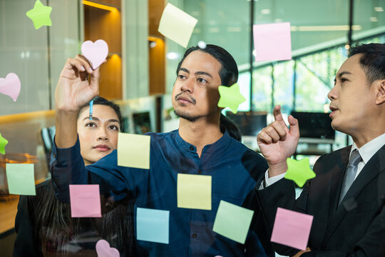 Businessman writing sticking adhesive notes on glass wall in office and team help to creative