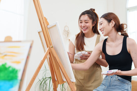 Two young beautiful, happy, cheerful Asian women smiling, laughing, painting watercolor together on canvas on wooden easel in cozy art studio at home with joy and fun