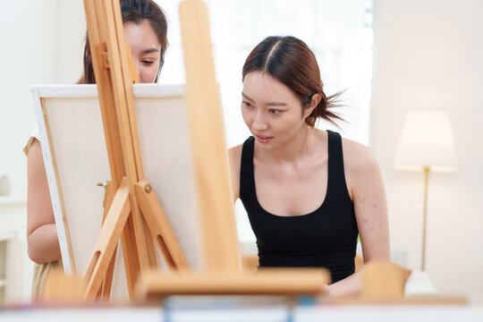 Young Asian Japanese woman artist in black tank top painting watercolor with paintbrush on canvas on wooden easel in white home art studio room with friends with calm and concentration