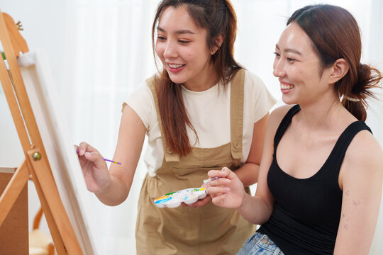 Two young beautiful, happy, cheerful Asian women smiling, laughing, painting watercolor together on canvas on wooden easel in cozy art studio at home with joy and fun