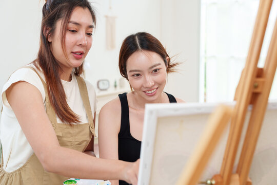 Two young beautiful, happy, cheerful Asian women smiling, laughing, painting watercolor together on canvas on wooden easel in cozy art studio at home with joy and fun. Friendship, mental wellbeing, di