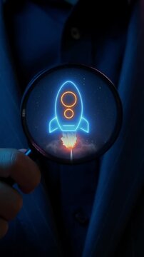Close-up vertical shot of hand holding magnifying glass to inspect glowing rocket launch target icon. Startup business growth acceleration and innovation launch strategy inspection concept.