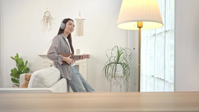 Young Asian woman playing a ukulele on the sofa during a relaxing weekend at home, enjoying music as a creative hobby