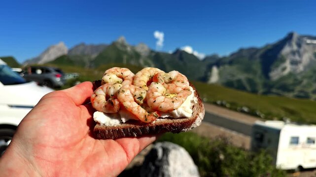 Hand holding shrimp sandwich against Pyrenees mountain landscape at Col d'Aubisque