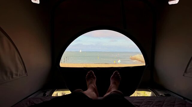 Man relaxing in a camper van roof tent enjoying the ocean view from his bed on the beach