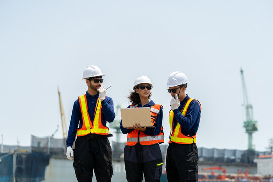 A group of engineers, wearing safety suits and helmets, are examining data on laptops in a shipyard and port area, with cranes and large ships in the background.