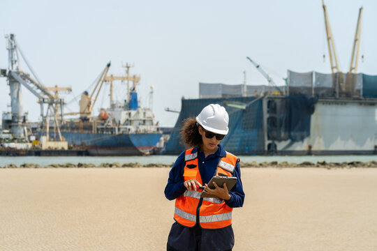 Portrait of female engineer in safety vest and helmet using radio and tablet at shipyard port, overseeing logistics and maritime shipping operations.