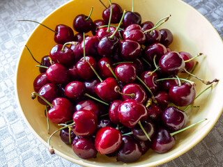 A close-up of fresh, ripe sweet cherries with stems in a yellow bowl. The glossy deep red fruits are naturally lit, highlighting their freshness and vibrant color © Gunars