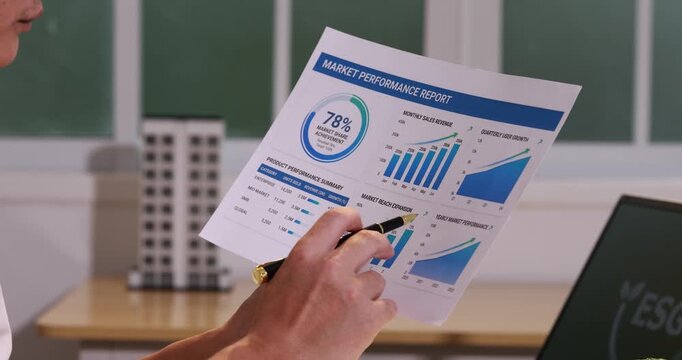 Person holding a business report with charts and graphs in an office, pointing at sales data with a pen