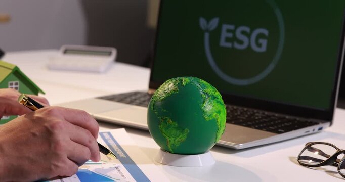 Hand working on an esg report beside a small green globe and laptop on a desk, business sustainability planning and environmental responsibility