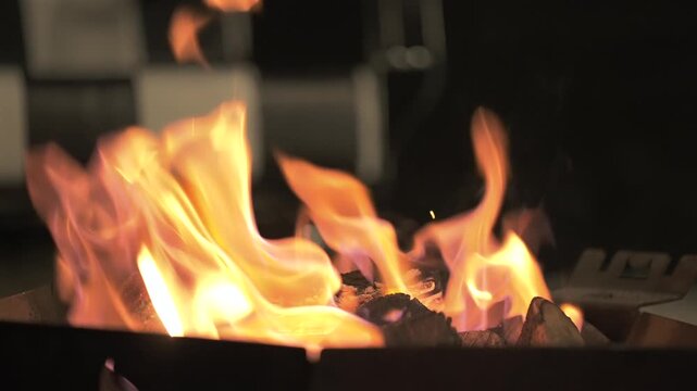 Intense orange and bright yellow flames roaring over burning logs on a rustic outdoor brazier at night. Static closeup shot.