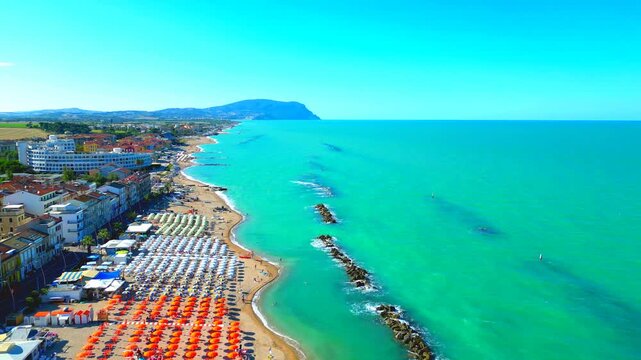 Stunning aerial truck left shot from Porto Recanati, azure waves of the Adriatic Sea washing the breakwaters and shore, a bird passing by towards the town, parasols, resorts, buildings, Mount Conero