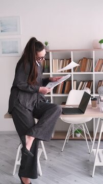 Vertical video. Business call. Office routine. Annoyed female manager talking on smartphone at desk with laptop analyzing paperwork at professional workplace.