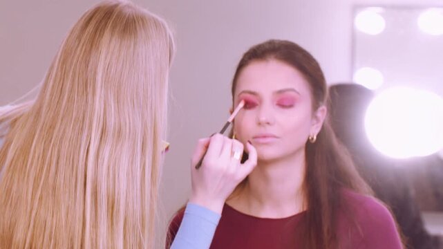 Color makeup. Professional visage. Female beautician applying eyeshadow to client eyelid using brush at workspace indoor.