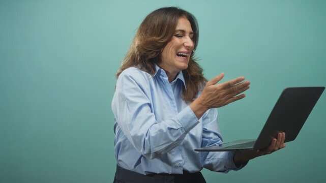 Middle age brunette woman holding laptop and blowing kiss with hand in studio teal backdrop; warm joy.