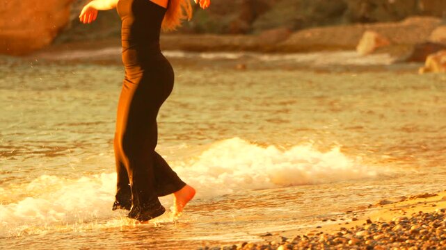 Beach sunset woman Slender woman in a black maxi dress walks gracefully along the shore at golden hour as waves lap her feet