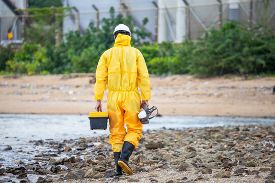 Environmental scientist in hazmat suit walking on beach near industrial plant, Worker in protective yellow suit holding toolbox and mask inspecting pollution and waste at coastal area