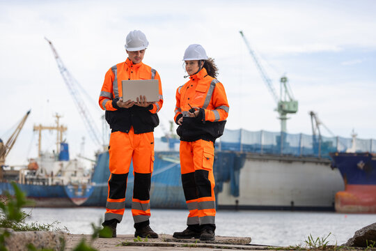 Logistic workers with laptop and radio standing at port shipping yard, Engineers in safety gear discussing cargo management by ship dock, Industrial worker team inspecting logistic operations outdoors