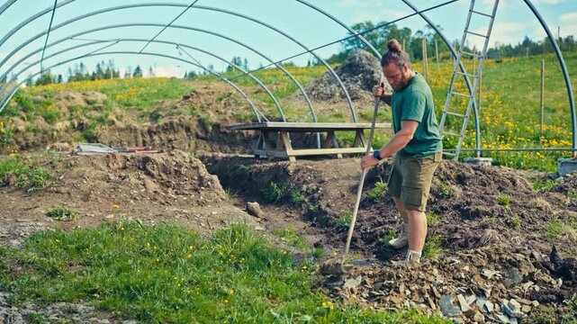 Man installing a greenhouse or polytunnel structure on a farm, showing agricultural work, construction materials, and the rural farming environment