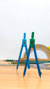 Two geometric compasses standing on a wooden desk with school supplies.