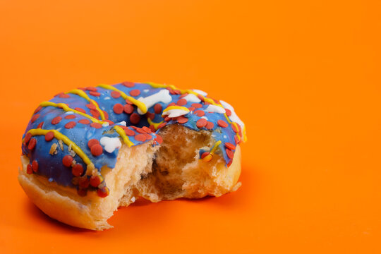 Bitten donut with vibrant blue icing and a mix of red, yellow, and white sprinkles, set against a bright orange background