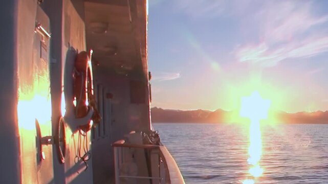 Witness a breathtaking Arctic Ocean sunrise from a moving ship. The golden sun casts a brilliant reflection on the shimmering waves, highlighting a peaceful maritime expedition as birds glide by.