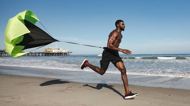 Athletic Black man running on beach with resistance parachute training