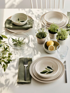 Fresh botanical kitchen table setting with ceramic plate, green napkin, herb plant, lemon, glassware, sunlight, and natural decor