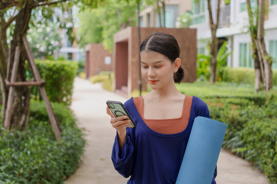 Asian Gen Z woman carrying yoga mat using smartphone while walking to urban park for wellness healthy lifestyle and smart city living.