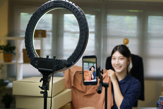 Excited young Asian woman live streaming a fashion clothing haul on smartphone with ring light, ideal for social media, e commerce, and influencer marketing concepts.