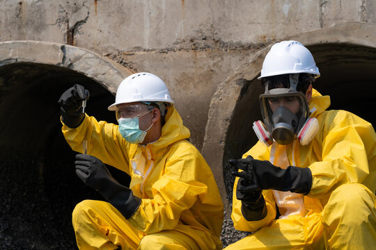Environmental engineer in yellow hazmat suit and helmet collecting water samples from a drainage pipe for pollution and environmental quality inspection.