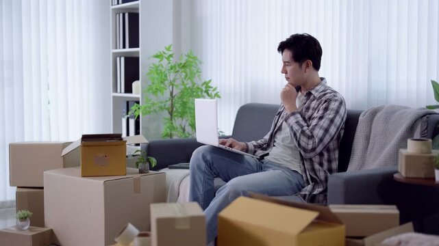 Young Asian man celebrating success while working on laptop surrounded by moving boxes in new apartment